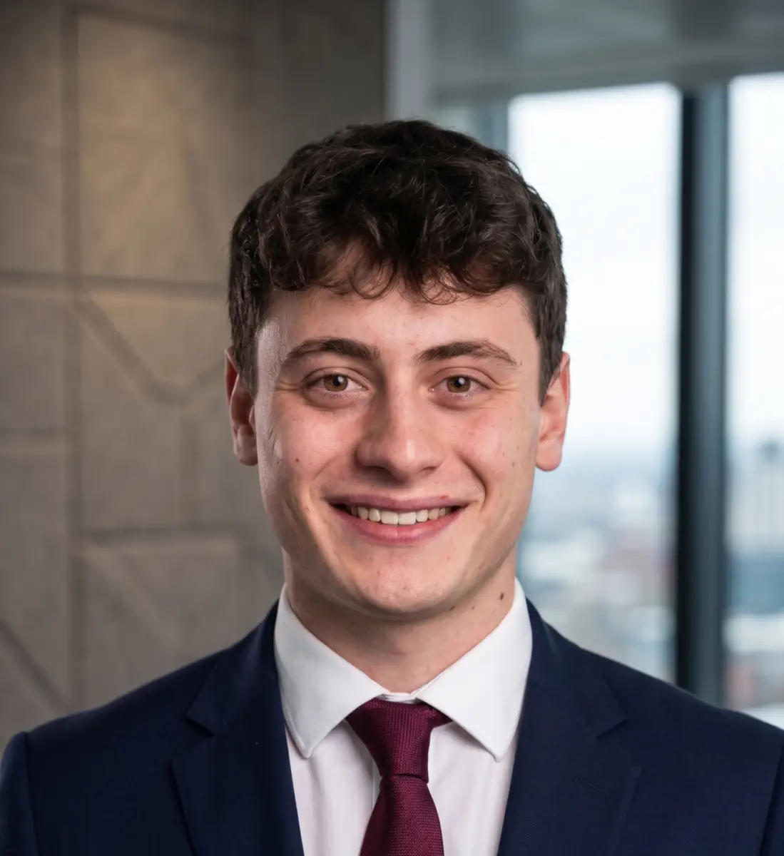 Close-up portrait of a smiling young man in a navy suit and burgundy tie, standing in a modern office setting.