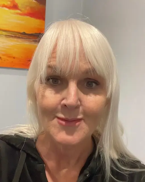 Portrait of a smiling woman with platinum blonde hair and bangs, indoors with a sunset painting on the wall behind her.